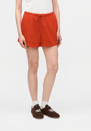 Osoba ubrana w jaskrawo pomarańczowe szorty z troczkami, białe skarpetki do kostek i ciemnobrązowe sznurowane tenisówki stojąca na jednolitym tle.