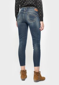 Jeans ajustados en denim azul oscuro con un acabado texturizado, que presentan una etiqueta de cuero marrón en la cinturilla y dos bolsillos traseros.