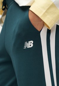 Χέρι στη τσέπη σκούρων πράσινων αθλητικών παντελονιών με λευκές κάθετες ρίγες και λευκό λογότυπο New Balance, φοράει κίτρινο και λευκό μακρυμάνικο πουκάμισο.