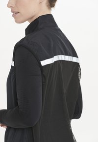 Svart väst med hög krage, med meshbak och reflekterande vit rand. Långa svarta ärmar med subtila mönster.