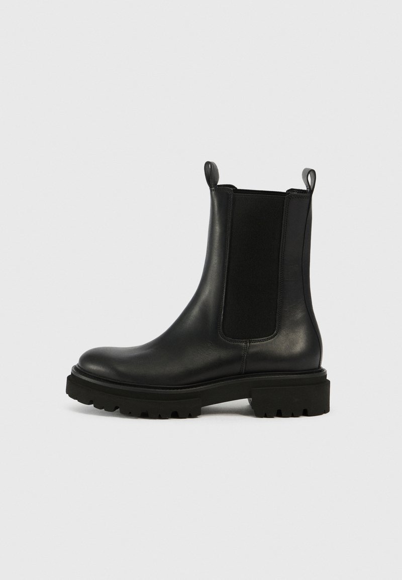 Bottines Chelsea en cuir noir avec panneau élastique sur les côtés, languettes de tirage, bout arrondi et semelle épaisse à relief, présentées sur un fond clair uni.