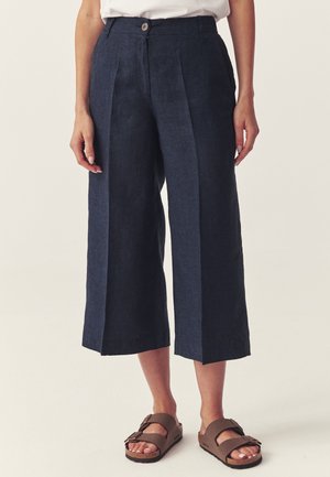 Kobieta w granatowych, szerokich, wysokich pantalonach z plisami z przodu i brązowych sandałach na podwójny pasek.