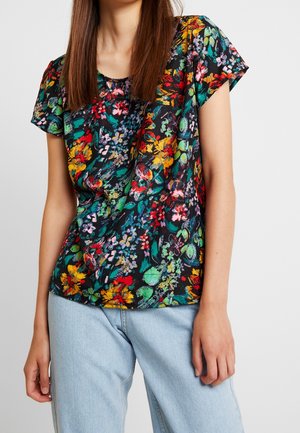 Vrouw draagt een zwart blouse met korte mouwen en levendige, meerkleurige bloemenpatronen, gecombineerd met lichtblauwe spijkerbroek.