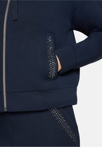 Navy sweatshirt with zipper, features a side pocket adorned with silver studs in a diagonal line, made from soft fabric with a smooth texture.