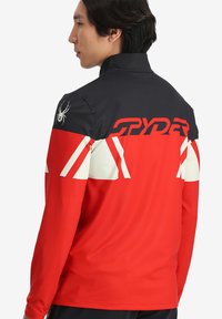 Giacca rossa e nera con colletto alto, dotata di un logo a forma di ragno, audace branding "SPYDER" e motivi triangolari bianchi sul retro.