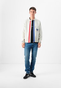 En ung mann står mot en hvit bakgrunn, iført en lys jakke med røde og blå striper, en svart t-skjorte, jeans og svarte sneakers.