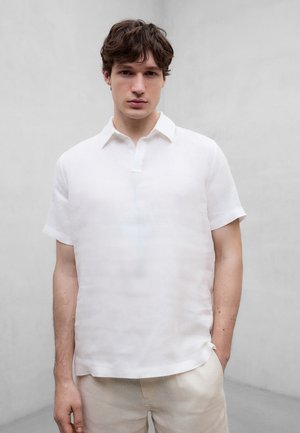 Joven con cabello castaño corto, vestido con una camisa blanca de manga corta con cuello y pantalones beige, sobre un fondo gris claro liso.