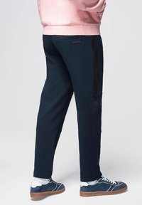 Námořnické tepláky s úzkým střihem, s boční kapsou a černým akcentním pruhem. Kombinované s modrými teniskami a bílými ponožkami.