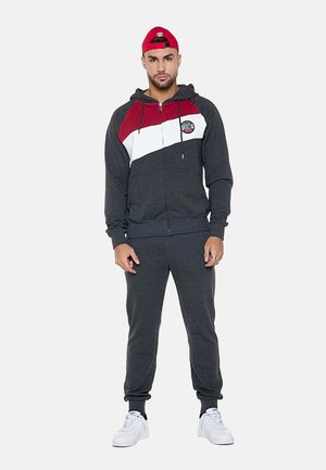 Uomo con berretto rosso, tuta grigio scuro con strisce diagonali rosse e bianche e scarpe da ginnastica bianche, in piedi davanti a uno sfondo bianco.