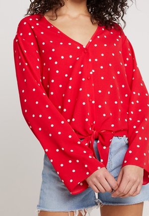 Blouse rouge à manches longues à pois blancs, nouée à la taille, associée à un short en denim bleu clair aux bords effilochés.