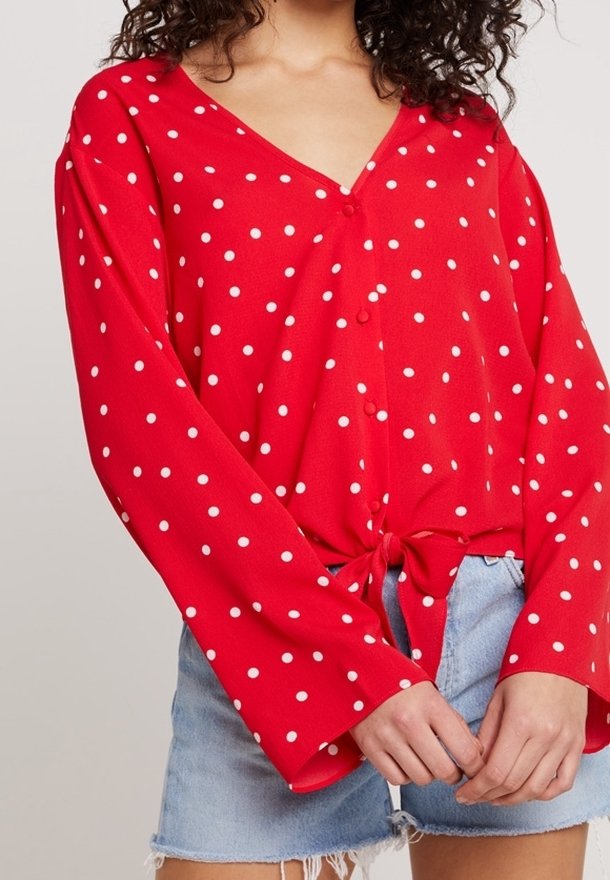 Blouse rouge à manches longues à pois blancs, nouée à la taille, associée à un short en denim bleu clair aux bords effilochés.
