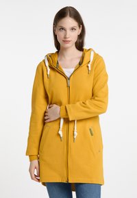 Felpa gialla con zip e cappuccio, cordini bianchi, tasche frontali e una texture morbida. Presenta un design casual con un taglio leggermente più lungo.