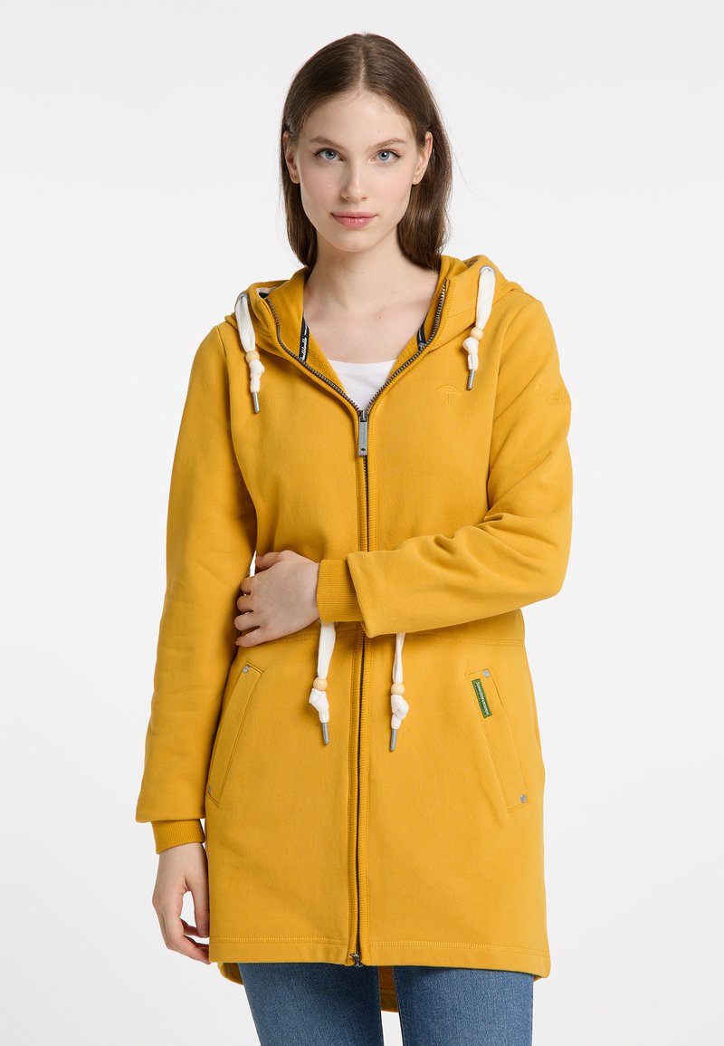 Felpa gialla con zip e cappuccio, cordini bianchi, tasche frontali e una texture morbida. Presenta un design casual con un taglio leggermente più lungo.