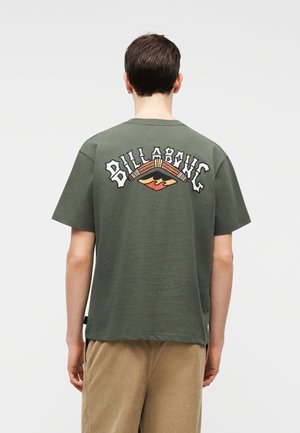 Person, der von hinten zu sehen ist, trägt ein olivgrünes Billabong-T-Shirt mit mehrfarbigem Logo und beige Hose vor einfarbigem Hintergrund.