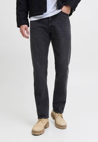 Hombre con jeans negros, botas de cordones beige, chaqueta negra y camiseta blanca de pie contra un fondo blanco liso.