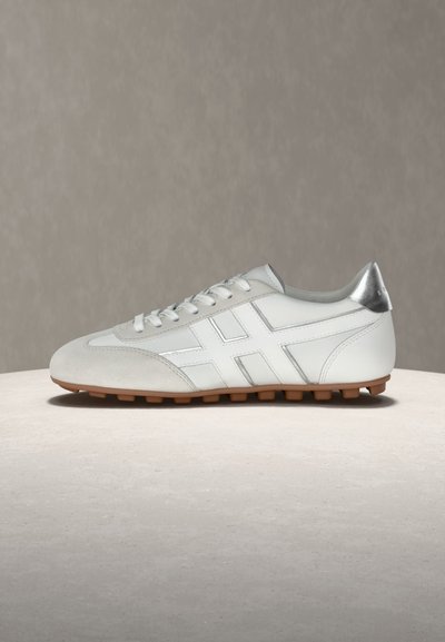 Λευκό δερμάτινο αθλητικό παπούτσι με ασημένια φτέρνα, μοτίβο λευκών σταυρωτών ραφών και καφέ λαστιχένια σόλα με καρφιά, εμφανιζόμενο από το πλάι πάνω σε φωτεινή επιφάνεια.