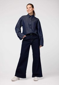 Camicia di jeans di colore blu scuro con bottoni frontali e maniche ampie. Jeans a vita alta e a gamba larga di colore scuro con cuciture a contrasto chiare. Sneakers bianche.