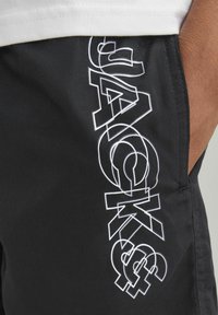 Pantalones cortos negros con un logo blanco que presenta la palabra "JACK". Diseño sencillo con una fuente angular, textura suave y detalle de bolsillo lateral.