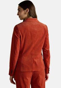 Sieviete, tērpusies rūsu krāsas korduroja žaketē un atbilstošajās biksēs, redzama no aizmugures uz vienkārša balta fona.