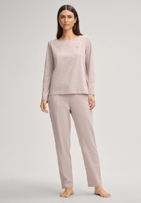 Vrouw staat blootvoets en draagt een bijpassende beige langemouwtop en broek met een klein herhalend patroon tegen een effen achtergrond.