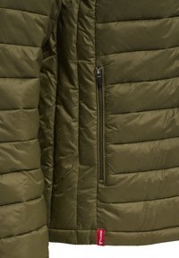 Olivgrüne gesteppte Jacke mit vertikaler Reißverschlusstasche vorne und einem kleinen roten Markenzeichen am unteren Saum.