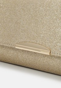 Złota kopertówka o brokatowej teksturze z zaokrąglonym złotym metalowym zapięciem, na białej powierzchni.
