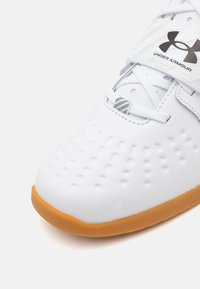 Hvid athletisk sko med tekstureret overflade og perforeringer. Har grå detaljer og en tan gummisål. Snor- og strop-lukningsdesign.