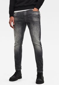 Vybledlé černé džíny s úzkým střihem, které mají odřeniny a mírný gradientní efekt. Stylizované s černými šněrovacími botami.