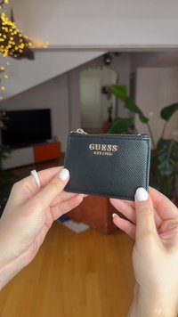 Portefeuille en cuir noir texturé avec un logo doré "GUESS EST 1981", fermeture éclair, et une forme rectangulaire compacte. Surface lisse et design minimaliste.