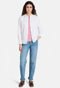 Witte blouse gedragen over een roze top, gecombineerd met lichtblauwe spijkerbroek en bruine schoenen. De outfit heeft een casual, ontspannen pasvorm.