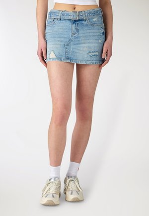 Donna che indossa una minigonna di jeans strappati azzurri, top corto bianco, calzini bianchi e sneakers color avorio, in piedi contro uno sfondo semplice.