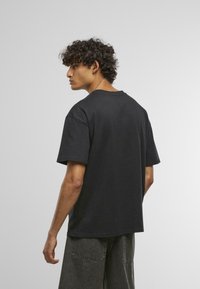 Sort bomulds T-shirt med korte ærmer og rund hals, vist fra bagsiden, parret med mørkegrå, løst siddende bukser.