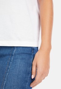 Weißes Baumwoll-T-Shirt mit lockerer Passform, kurzen Ärmeln und einem dezenten Logo am Saum. Getragen mit blauen Denim-Jeans, die eine gepflegte Hand zeigen.