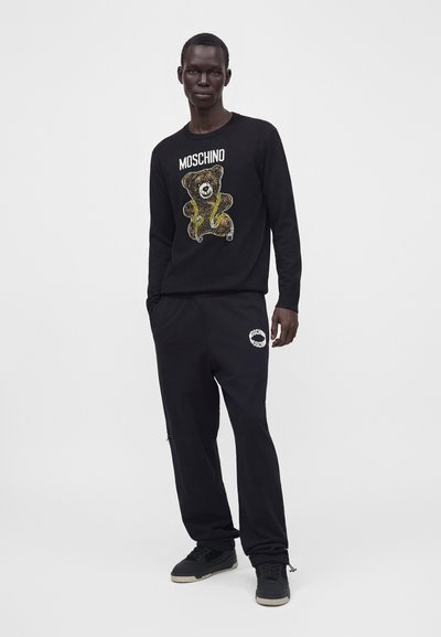 Homme portant un sweat Moschino noir avec un motif d'ours en peluche et un pantalon noir assorti avec un logo sur la cuisse, debout sur un fond blanc uni.