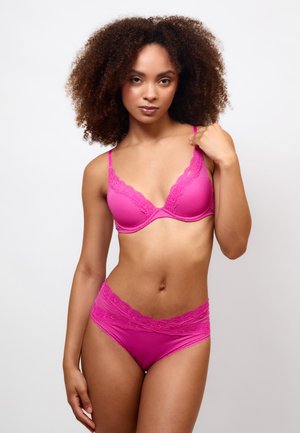 Femme aux cheveux bouclés portant un soutien-gorge rose vif bordé de dentelle et une culotte taille haute assortie sur un fond blanc uni.