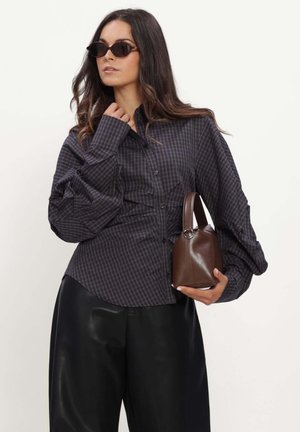 Femme portant un chemisier à carreaux sombre et un pantalon noir, tenant un sac à main marron et portant des lunettes de soleil, debout devant un fond uni.