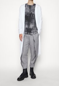 Cardigan bianco a maniche lunghe sopra una maglietta grigia a tie-dye, abbinato a pantaloni grigi plissettati e stivali neri con lacci.