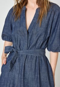 Abito in denim blu con maniche a sbuffo corte, chiusura con bottoni frontale e cintura da annodare in vita. Presenta una vestibilità comoda e una texture naturale.