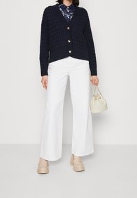 Marinblått stickat kardigan med ett texturerat mönster, guldknappar, i kombination med vita vida byxor och beige plattformsskor, hållande en liten krämfärgad väska.