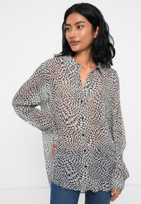 Black and white patterned sheer button-up shirt with long sleeves, collar, and relaxed fit, worn with blue jeans.