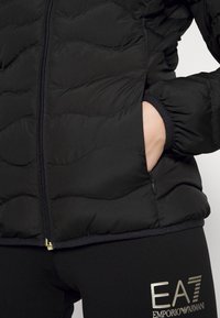 Czarna pikowana kurtka z falistym przeszyciem, zamkiem błyskawicznym i elastycznymi mankietami. Posiada kieszenie oraz złoty akcent przy krawędzi.