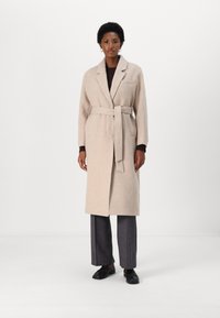 Cappotto di lana beige con vita cinturata, colletto a scollo e tasche laterali, indossato sopra un maglione scuro, abbinato a pantaloni grigi e scarpe nere.