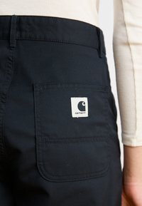 Black Carhartt pants with a rear patch pocket, featuring a white label with the brand logo. Material appears durable with a smooth texture.
