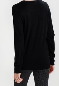 Schwarzer langärmeliger Pullover mit rundem Ausschnitt, mit glatter Textur und geripptem Saum, kombiniert mit gepunkteten schwarz-weißen Hosen.