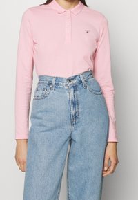 Långärmad rosa polo med krage och knappslå, ihop med ljusblå högmidjade jeans. GANT-logotyp på bröstet.