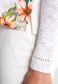 Weiße gestrickte Ärmel neben weißen Hosen mit einem floralen Stoffetikett, das orange und grüne Blumen zeigt und das Label "Napapijri" trägt.