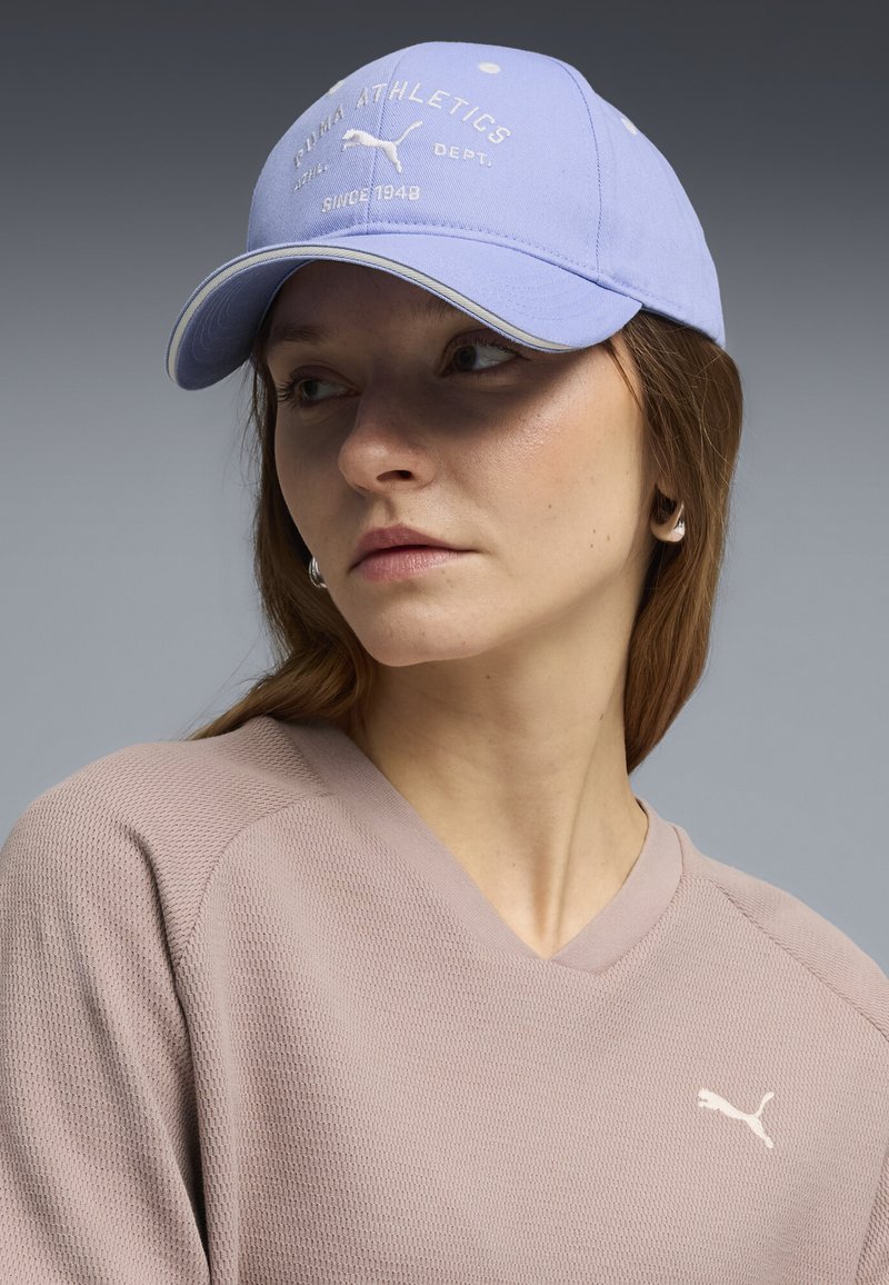 Femme portant une casquette Puma lilas clair avec texte et logo, ainsi qu'un haut à manches longues texturé assorti avec le logo Puma, regardant sur le côté.