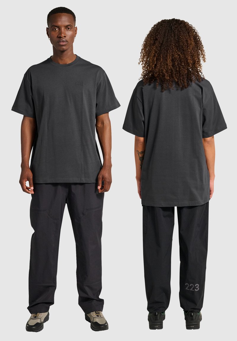 Dunkelgraues, oversized T-Shirt mit rundem Ausschnitt, aus Baumwolle. Getragen mit lockeren schwarzen Hosen, die an einem Bein die sichtbare Nummer "223" haben.