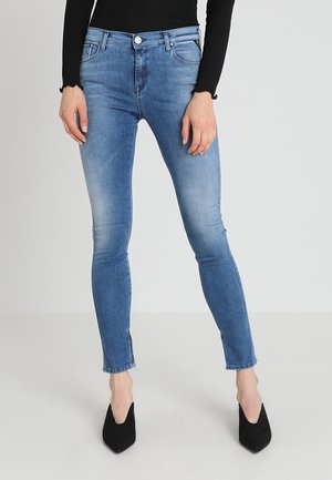 Kobieta w jasnoniebieskich obcisłych dżinsach z zamkami przy kostkach, w połączeniu z czarnymi butami na szpic i czarną bluzką z długim rękawem.