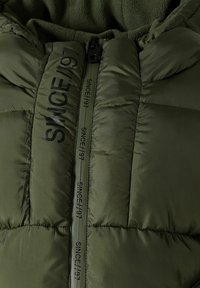 Giacca puffer verde oliva con design trapuntato, dotata di una cerniera nera e testo in rilievo "SINCE/97" sul fronte e sul lato. Fodera in morbido pile.